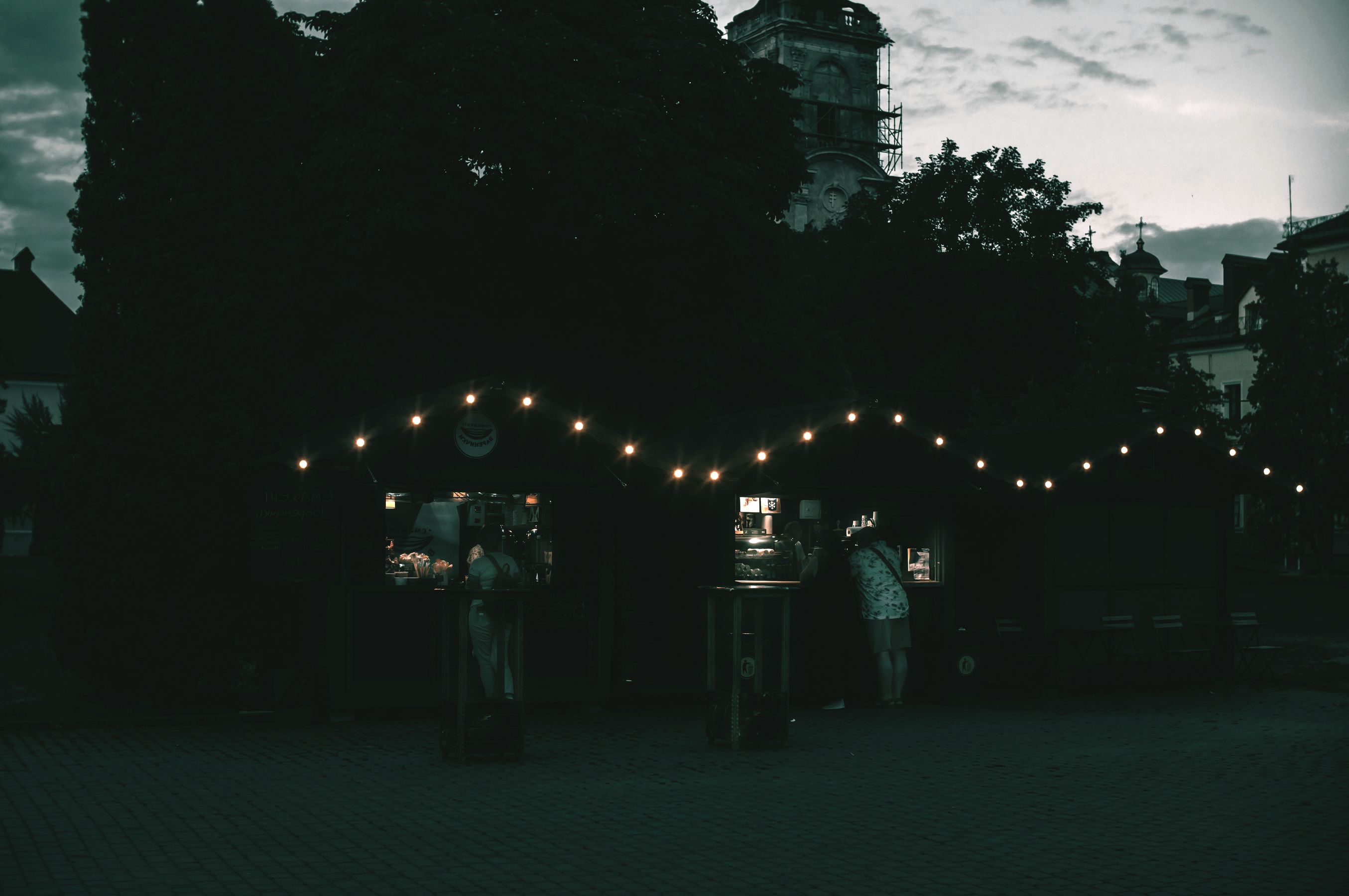 Twilight urban market stalls with dim, moody lights — the night the maker sets up the glowing brain and the case of forty bottles.